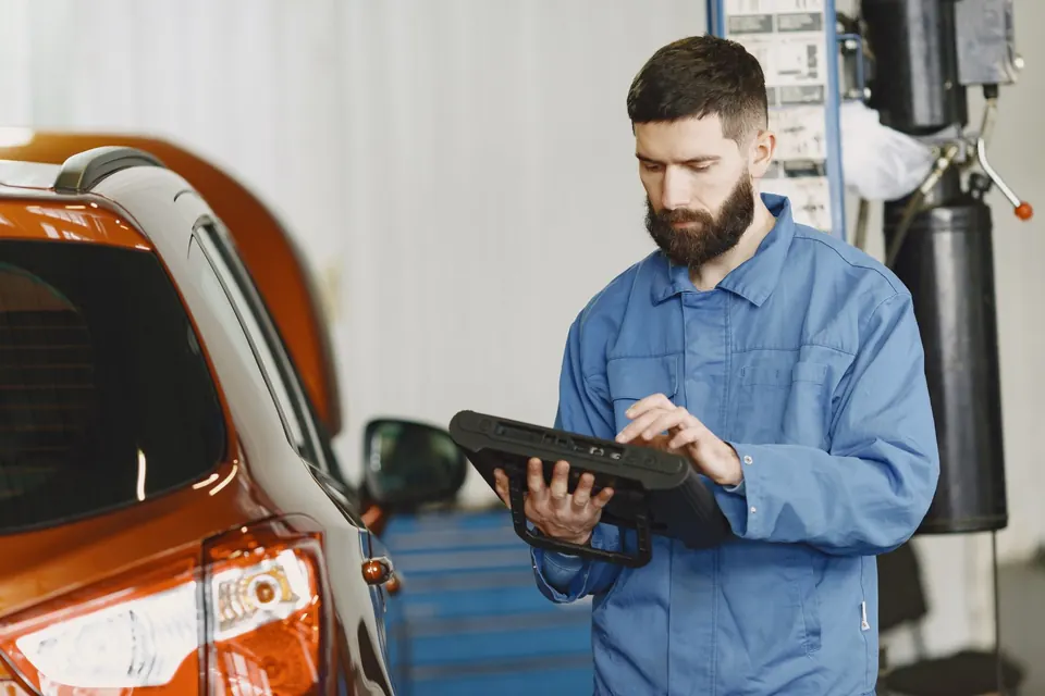 A technician in coveralls reading a diagnostic tablet beside a car in a service bay